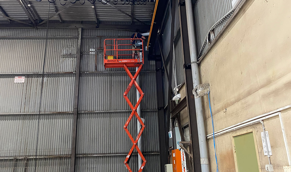 An electrician in an orange scissor lift working on pipes in ceiling